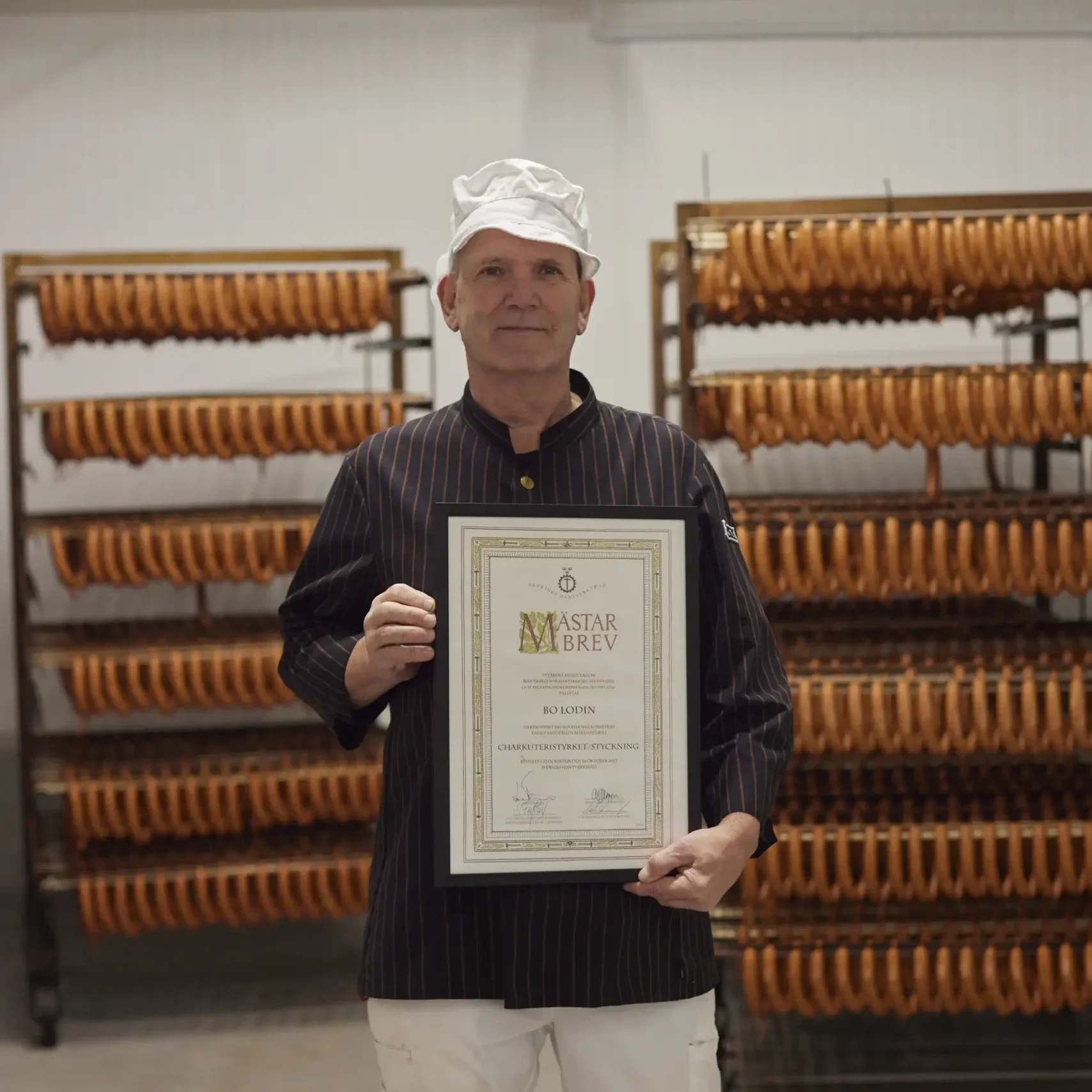 En korvarbetare i vit hatt håller upp en tavla med texten "Mästarbrev" tydligt synligt. I bakgrunden står hyllplan med hängande korvar.